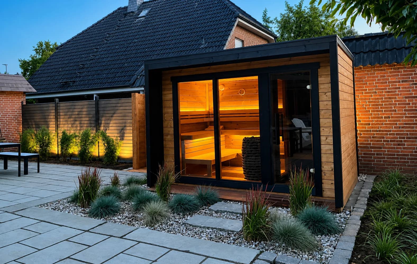 Moderne Outdoor-Gartensauna mit Glasfront und warmer Innenbeleuchtung in einem Wellnessgarten mit Ziergräsern in Elmshorn